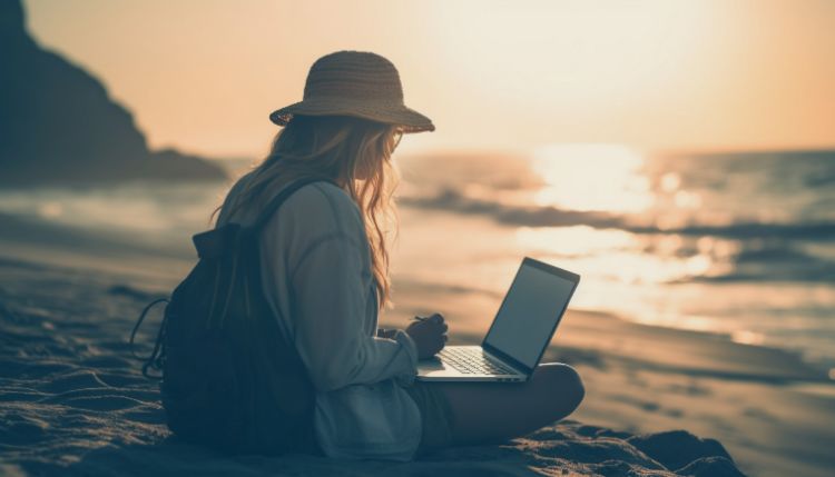 woman with long hair and wearing a hat sitting on a beach at dusk working on a her laptop